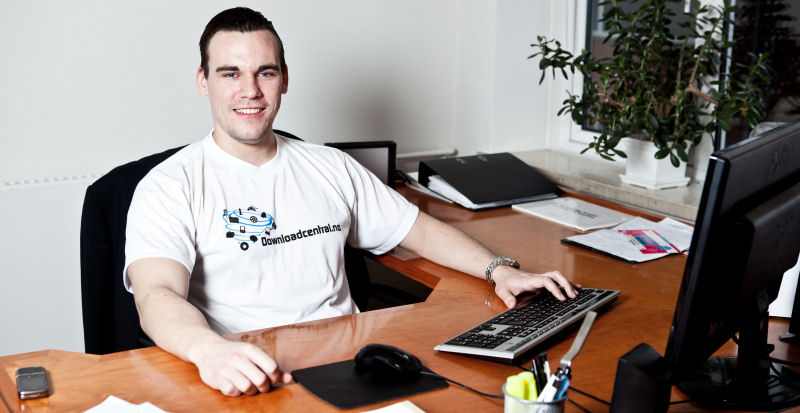 Grunnleggeren av Downloadcentral.no sitter ved skrivebordet på kontoret, arbeider ved datamaskinen og bruker en T-shirt med Downloadcentral-logo.
